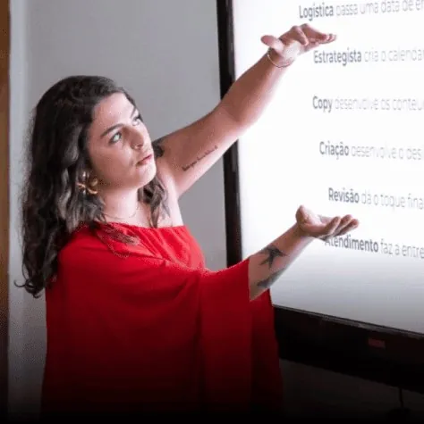 Stefania dando aula no palco, de vestido vermelho, apontando para a tela